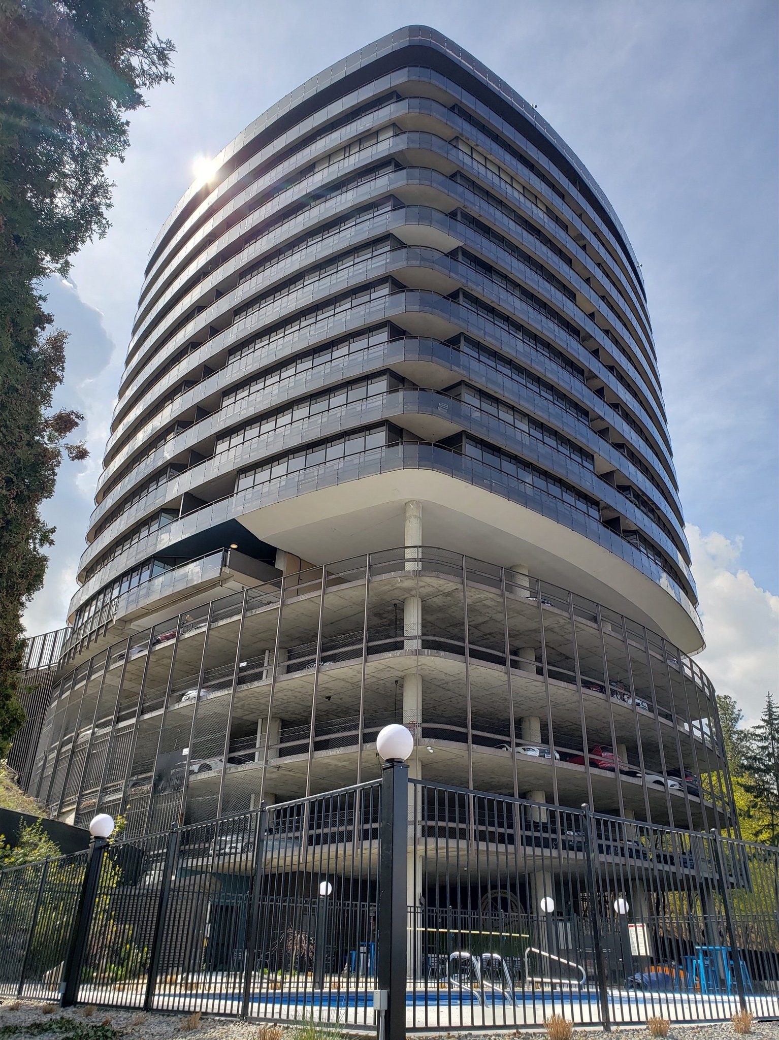 A tall, multi-story modern building with glass exterior and an open ground level, surrounded by a black metal fence with white spherical lights. Trees and blue sky are in the background.